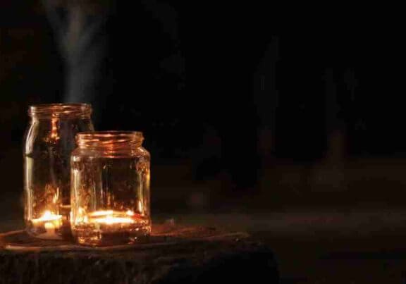 Two jars with lit tea candles in them sitting on a wood stump at night.